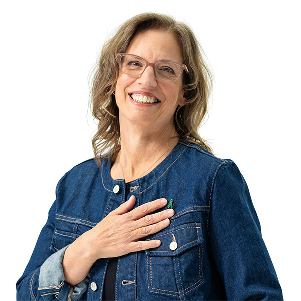 Kathleen, wearing a jean jacket and glasses, with her right hand over her heart, looking at the camera, smiling