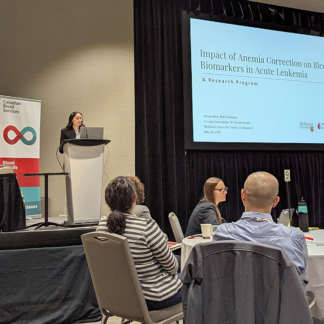 Dr. Dimpy Modi standing at a podium in front of a Canadian Blood Services backdrop, presenting her research on a large screen.
