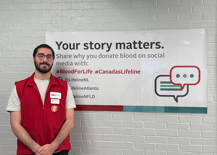 A young man wearing a red volunteer vest smiles in front of a Canadian Blood Services poster, with the words “Your story matters” printed on it.