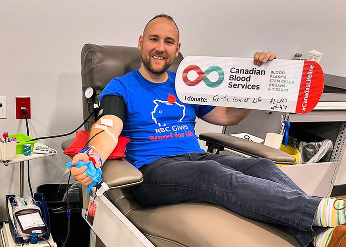 A man wearing a blue shirt donating blood in a donor centre, holding a sign that reads “For the love of life… always. #47”