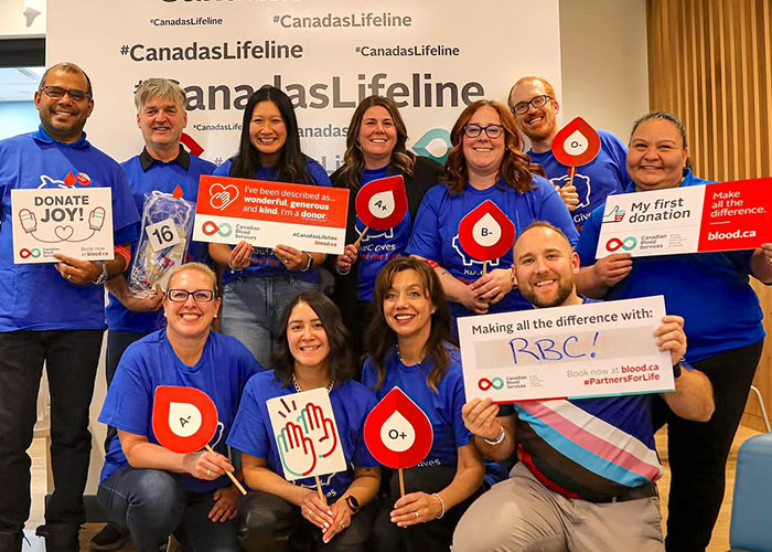 A group of people stand in front of a selfie wall printed with #CanadasLifeline holding up signs and celebrating their donations.