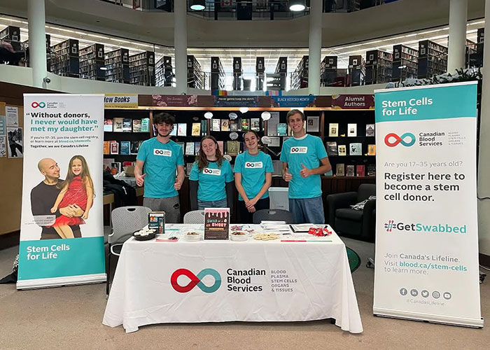Four students wearing teal t-shirts stand at a Canadian Blood Services table, flanked by pop-up banners about stem cells.