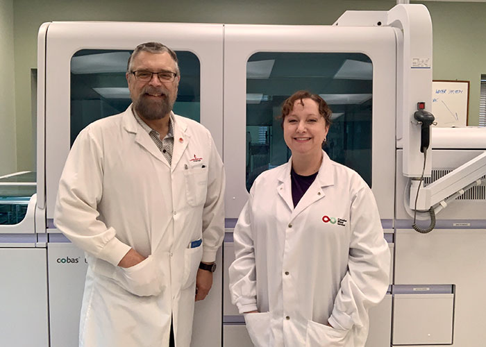 Douglas Ems & Valerie Conrod in front of a large machine wearing white lab coats