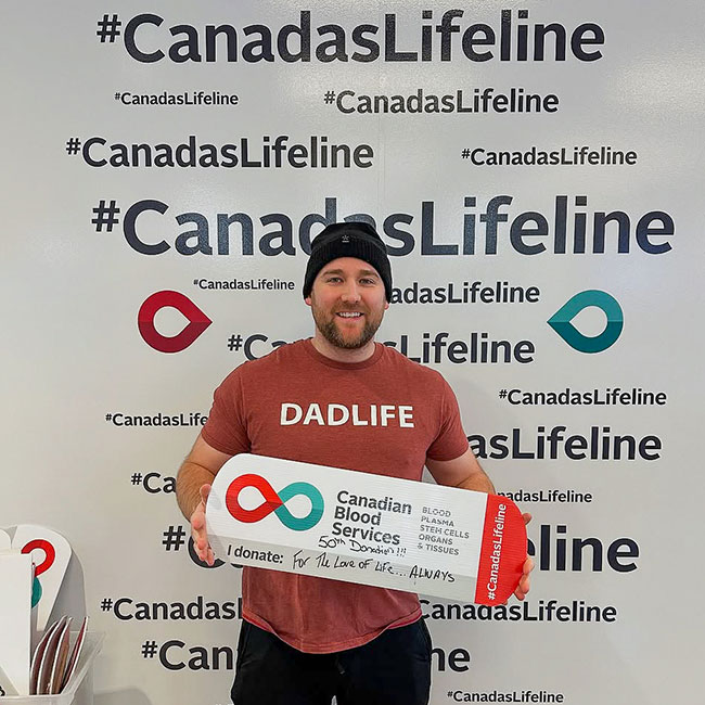 A smiling man with a red shirt holding a Canadian Blood Services sign that reads “50th donation! For the love of life… Always”