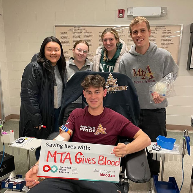 A young man holds a sign that reads “MTA Gives Blood” while donating blood. A group of four students wearing collegiate sweatshirts stand behind him.