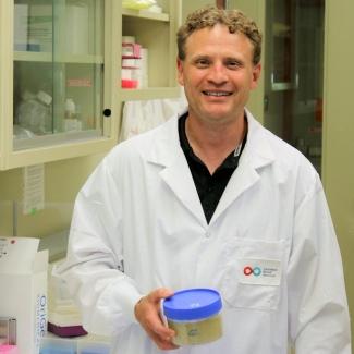 Dr Nicolas Pineault wears a lab coat and holds a blue plastic container in a stem cell lab