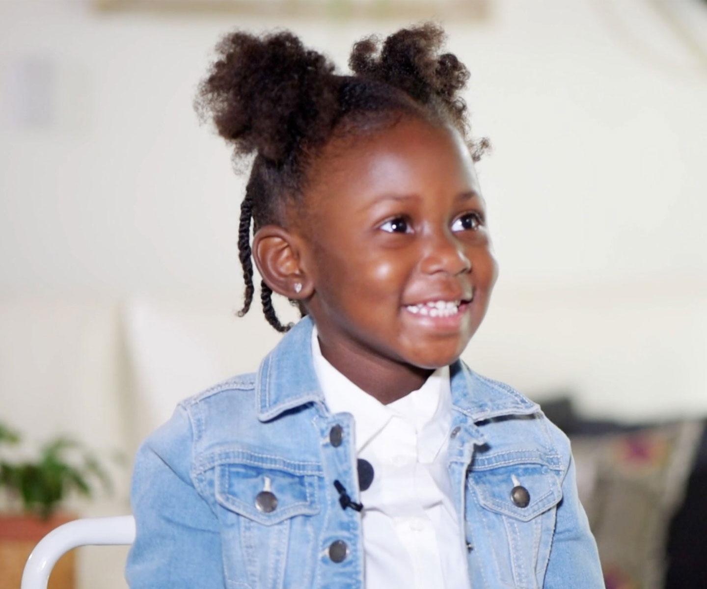 Preschool age girl in a jean jacket, seated indoors, looking off-camera and smiling broadly
