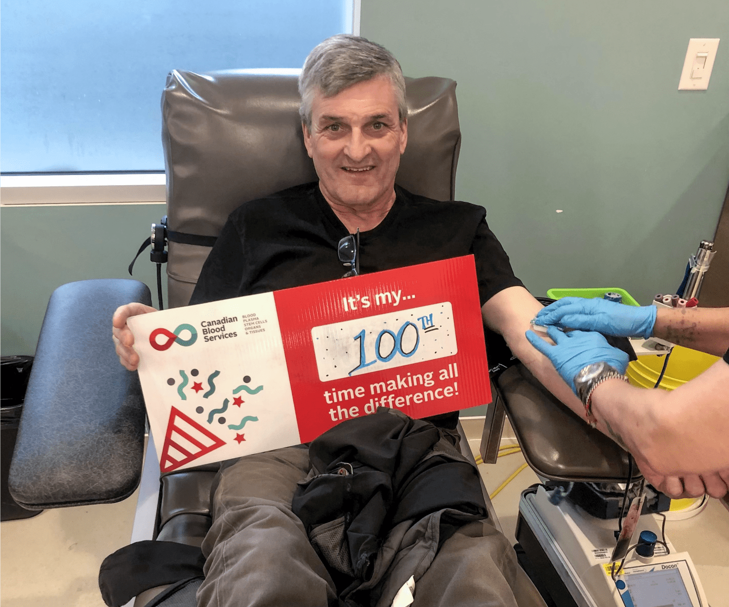 Blood donor in chair with sign that says "It's my 100th time making all the difference"