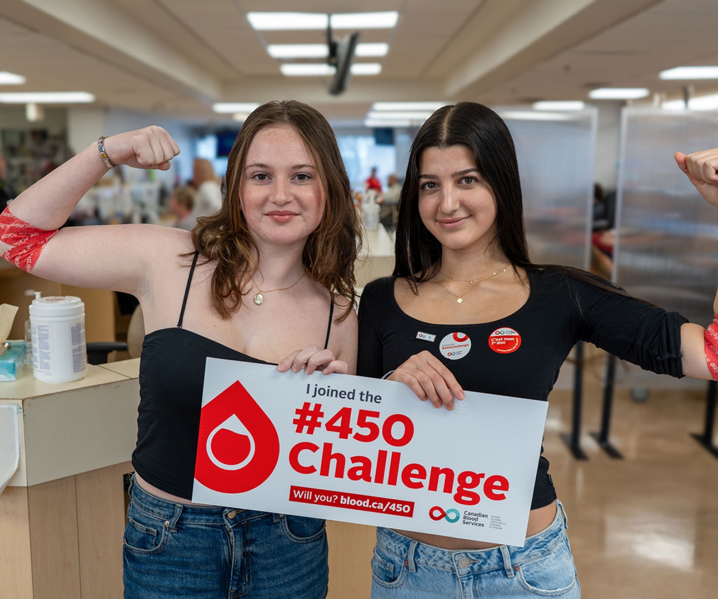 Two first-time blood donors in a donor centre flexing arms with pressure bandages and holding a sign that says “I joined the 450 Challenge”