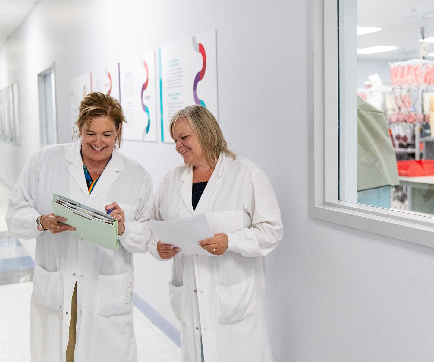 Two people wearing white lab coats standing in hallway at Concourse Gate facility looking at a document together. Blood hanging from bags in processing facility is visible in background. 