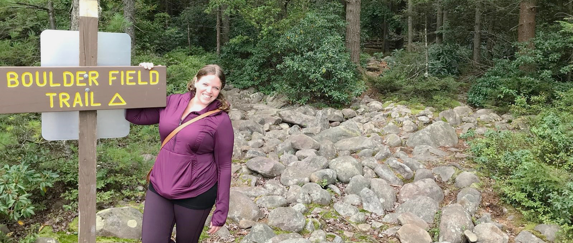 A woman stands on a trail of small boulders with a dense forest of green trees in back. 