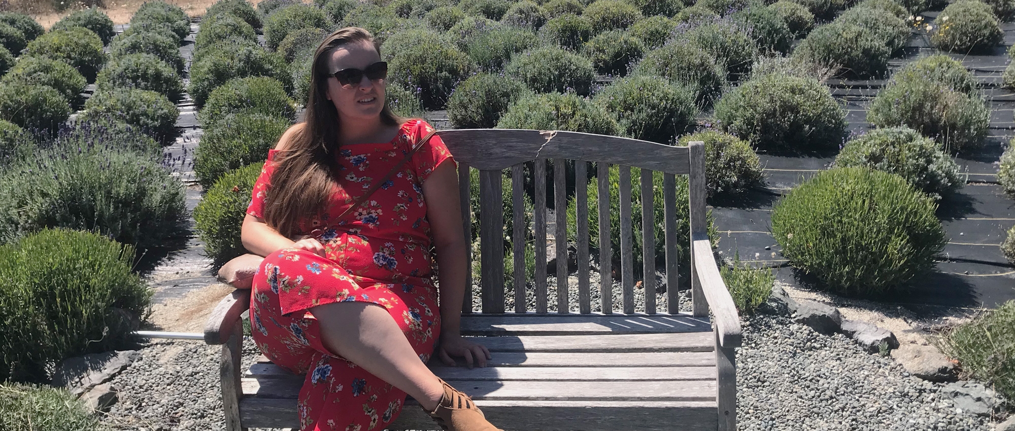 Woman in sunglasses sits on a bench in front of a greenhouse and rows of bushes