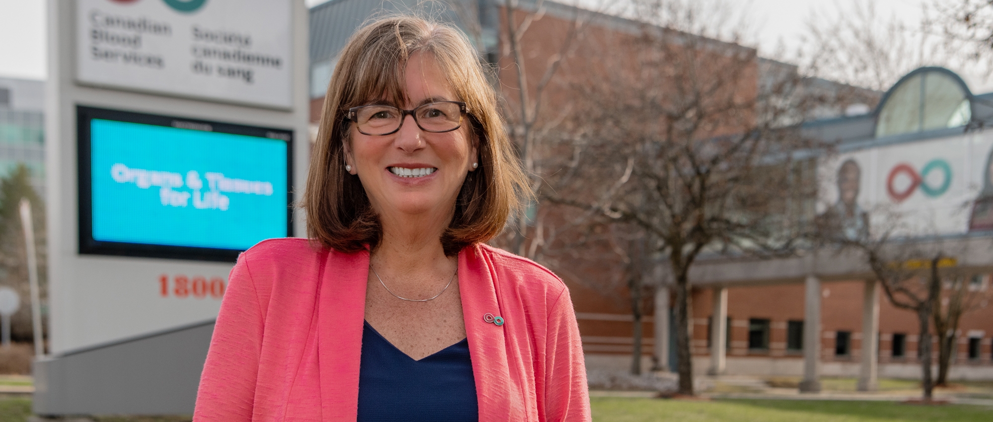 Image of Margaret Miedema, director of philanthropy standing outside of Canadian Blood Services head office's front entrance in front of the signage