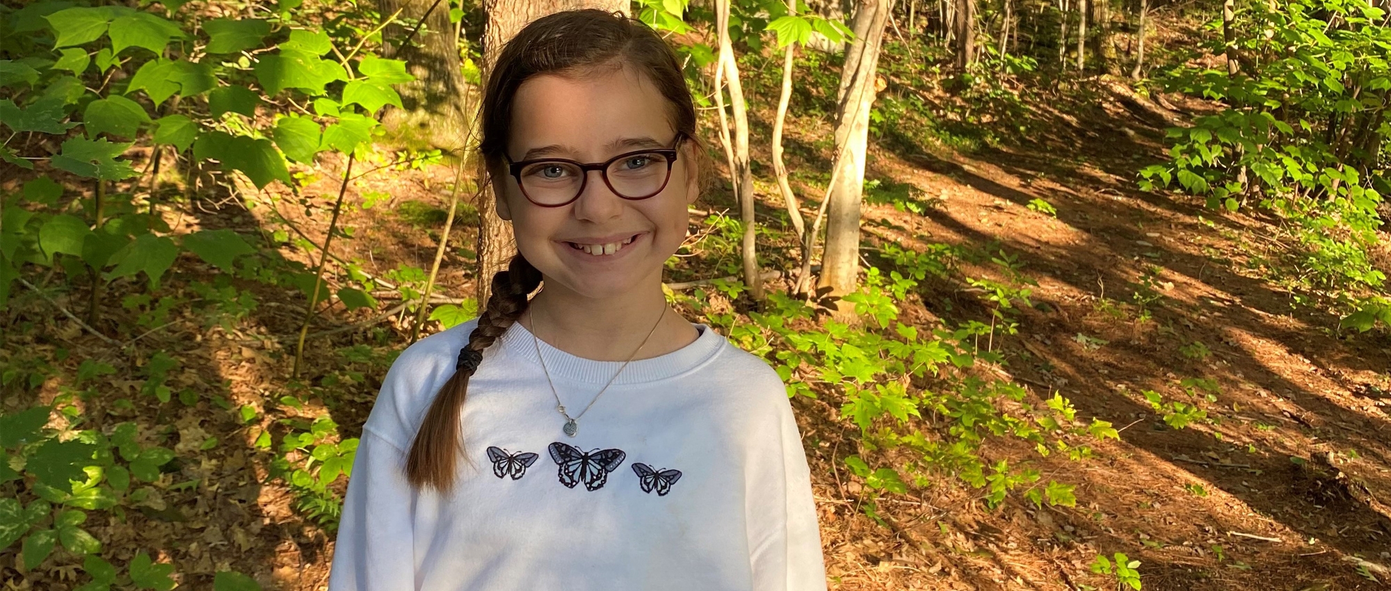 A young teen stands among trees, smiling at the camera