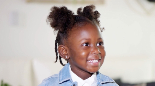 Petite fille d’âge préscolaire en veste en jean, assise en intérieur, regard hors champ et souriante