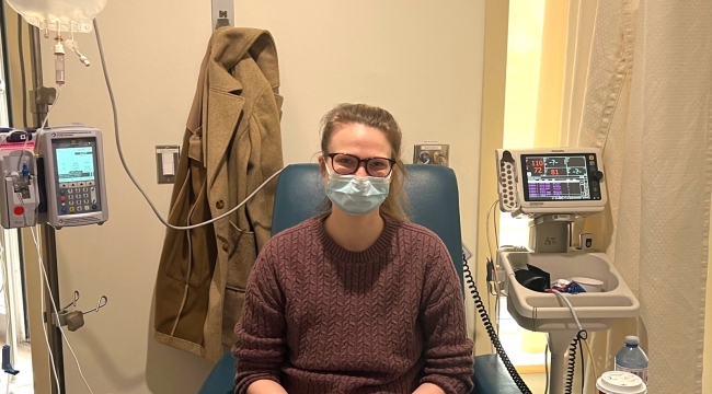 Masked person sits in blue hospital chair with intravenous tubes in one arm and a small knitting project in lap. A large bag of immunoglobulin being infused hangs above.  