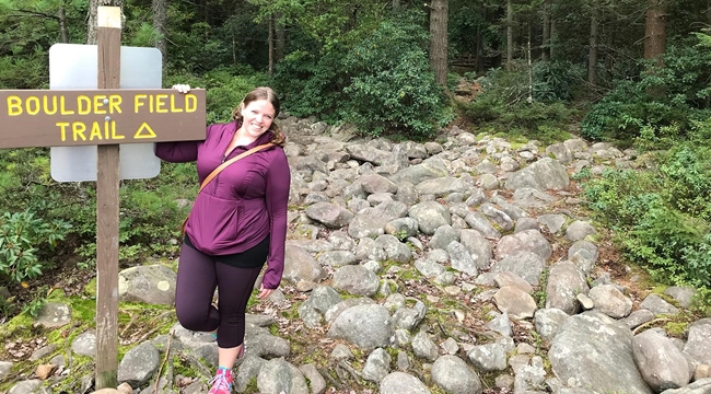 A woman stands on a trail of small boulders with a dense forest of green trees in back.