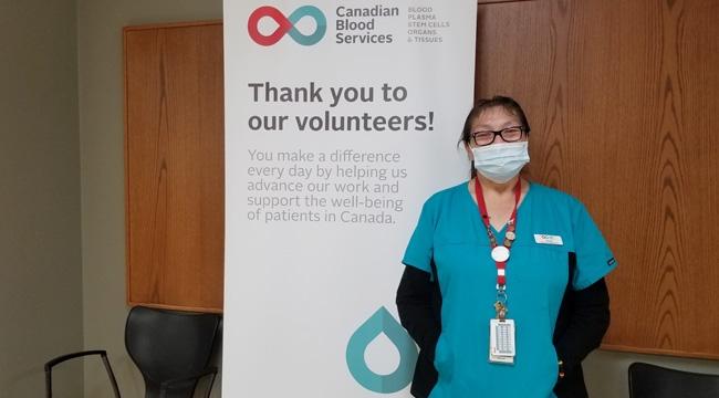 Donor services representative Annette Regalado, dressed in black and blue scrubs, stands in front of a Canadian Blood Services banner which reads: “Thank you to our volunteers.”