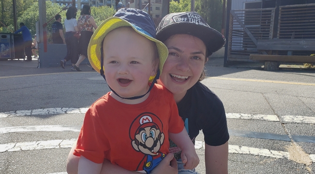 Mother crouched down on paved road with arm around the waist of her 2-year-old son and passersby in the background