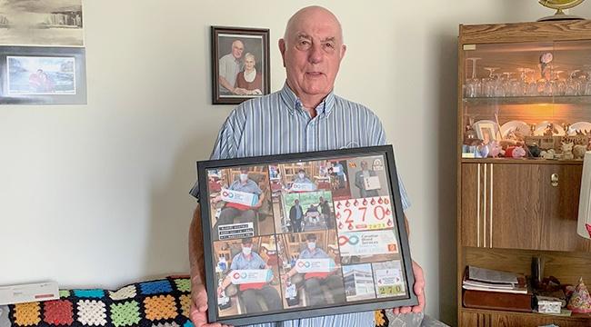 An 87-year-old man in a striped shirt holds a frame filled with mementoes of his blood donations.