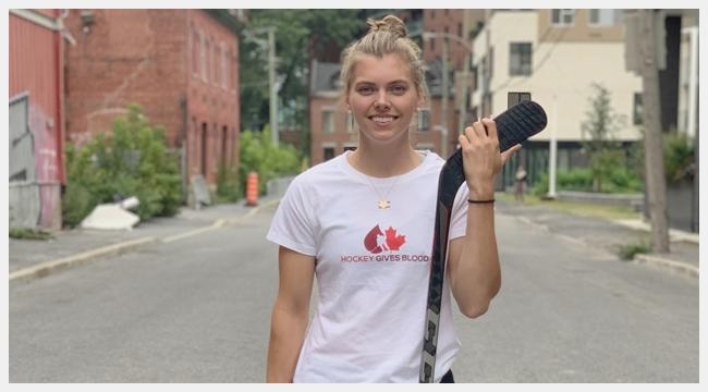 Image of a white blonde girl wearing a Hockey Gives Blood t-shirt and holding a hockey stick.