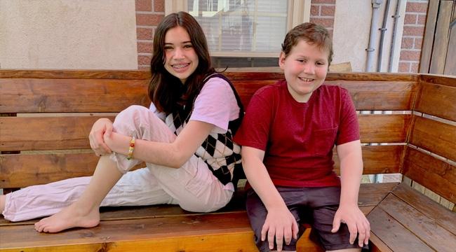 A sister and brother sit together on a bench outside their home.