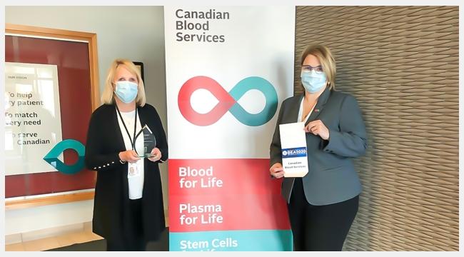 Image of director of testing Nancy Angus and director of supply chain operations Andrea Moore holding the award for best organization in Brampton’s life sciences sector standing in front of the Canadian Blood Services pull up stand.