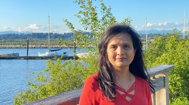 Canadian Blood Services donor care associate in B.C.’s s lower mainland stands on a wooden deck by a lake with green plants and boats behind her. 