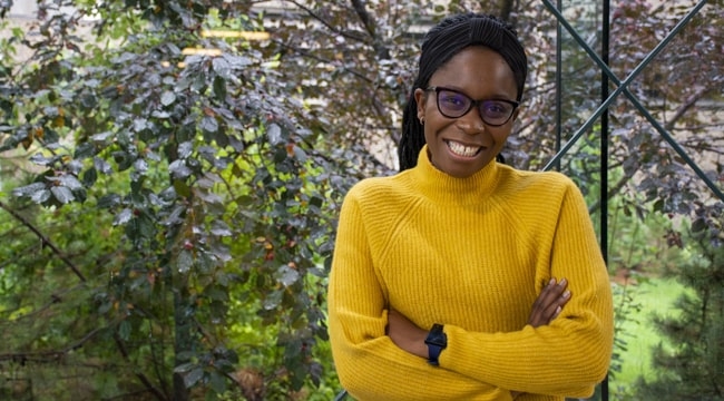 A woman wearing a yellow sweater and black-rimmed glasses with long black hair smiles with her arms crossed standing in front of leafy greenery outdoors on a sunny day.