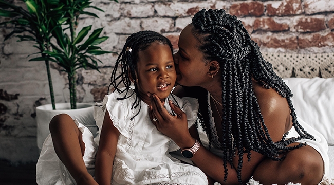 A mother gives her young daughter a kiss on the cheek, sitting on a bed in front of a brick wall. 